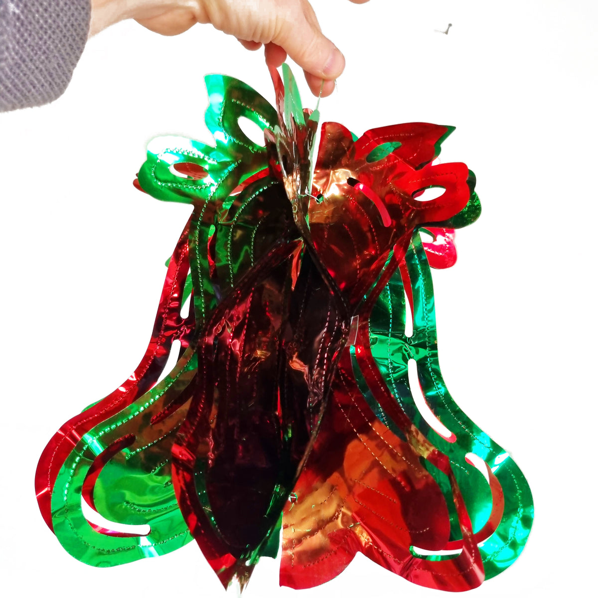A hand holding a shiny red and green Christmas ornament shaped like a stylized reindeer head with layered metallic bell-like shapes, shown against a plain white background.