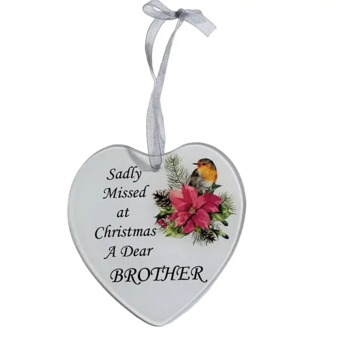 A heart-shaped white Christmas ornament hanging on a decorated tree. The ornament has a gray ribbon loop and reads 'Sadly Missed at Christmas A Dear BROTHER' with an illustration of red flowers and a small bird.