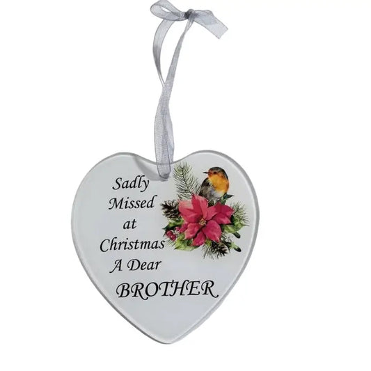 A heart-shaped white Christmas ornament hanging on a decorated tree. The ornament has a gray ribbon loop and reads 'Sadly Missed at Christmas A Dear BROTHER' with an illustration of red flowers and a small bird.