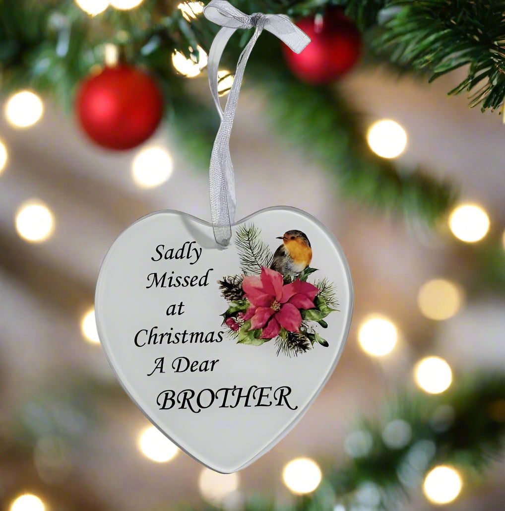 A heart-shaped white Christmas ornament hanging from a silver ribbon on a Christmas tree branch. The ornament features the printed message 'Sadly Missed at Christmas A Dear BROTHER' alongside an illustration of a robin bird and a pink flower, with a warm bokeh background of Christmas lights.