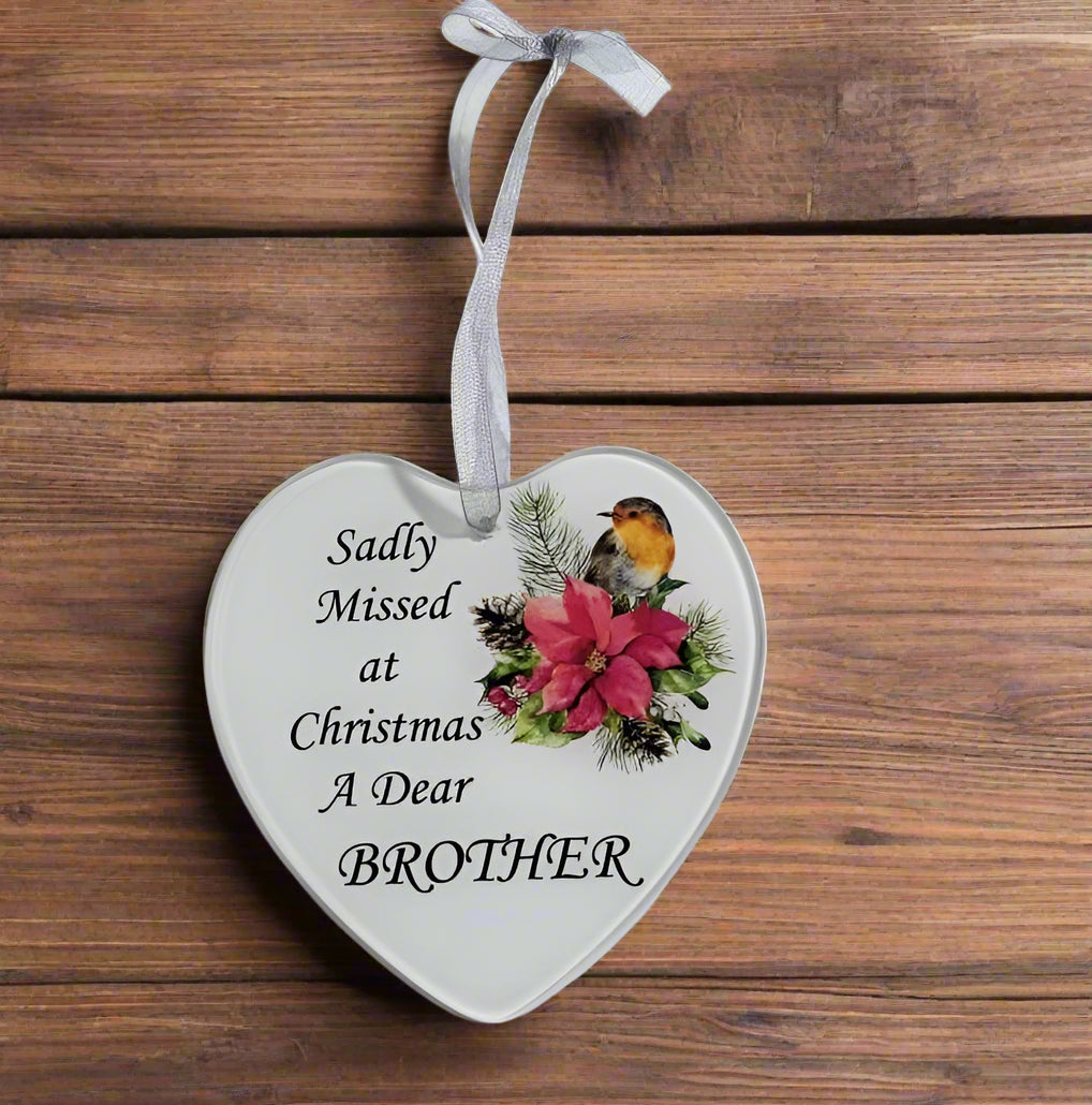 A heart-shaped white ceramic Christmas ornament with a silver hanging ribbon, featuring the printed text 'Sadly Missed at Christmas A Dear BROTHER' alongside a festive illustration of a robin bird and a pink poinsettia, displayed on a rustic wooden background.