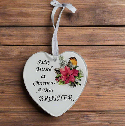 A heart-shaped white ceramic Christmas ornament with a silver hanging ribbon, featuring the printed text 'Sadly Missed at Christmas A Dear BROTHER' alongside a festive illustration of a robin bird and a pink poinsettia, displayed on a rustic wooden background.