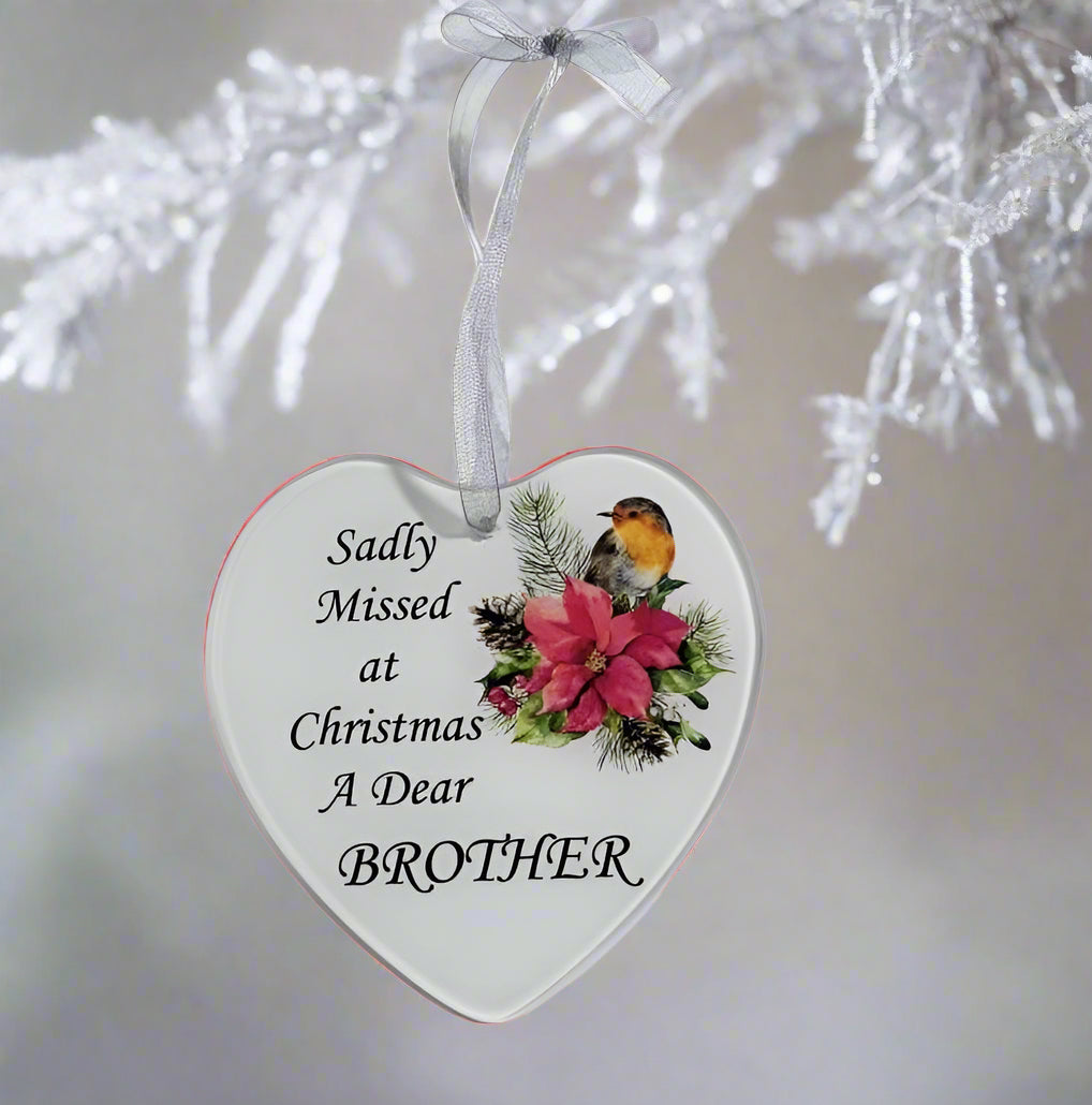 A heart-shaped white Christmas ornament hanging from a white ribbon, printed with the text 'Sadly Missed at Christmas A Dear BROTHER' and decorated with a robin bird and pink flowers, set against a blurred white Christmas tree background.