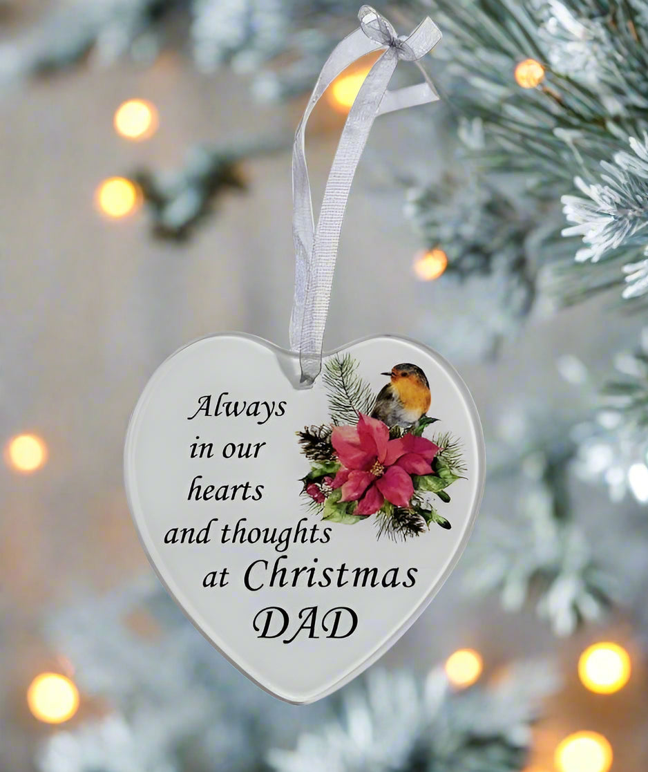 A heart-shaped white Christmas ornament hanging from a silver ribbon, printed with the message 'Always in our hearts and thoughts at Christmas DAD' alongside a robin bird and pink flowers, set against a blurred Christmas tree background with warm bokeh lights.