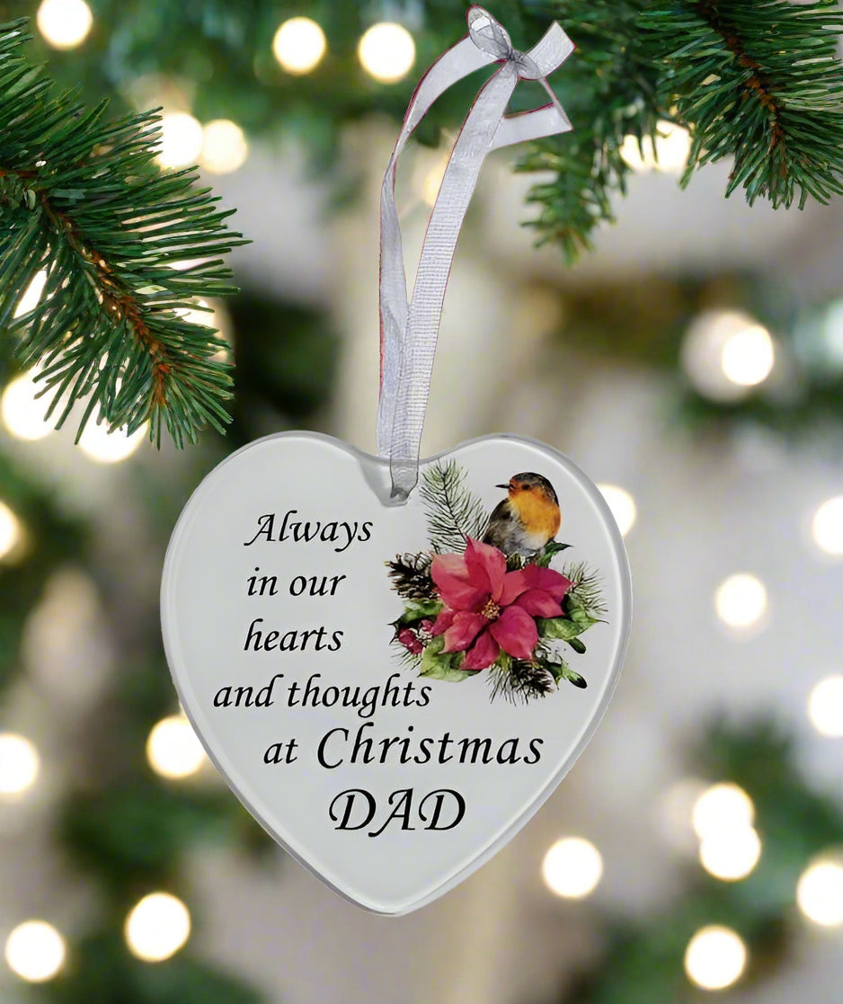 A heart-shaped Christmas ornament hanging from a white ribbon, featuring the text 'Always in our hearts and thoughts at Christmas DAD' alongside an illustration of a bird and pink flowers. The ornament is shown hanging from pine branches with a warm, blurred background of glowing Christmas lights.