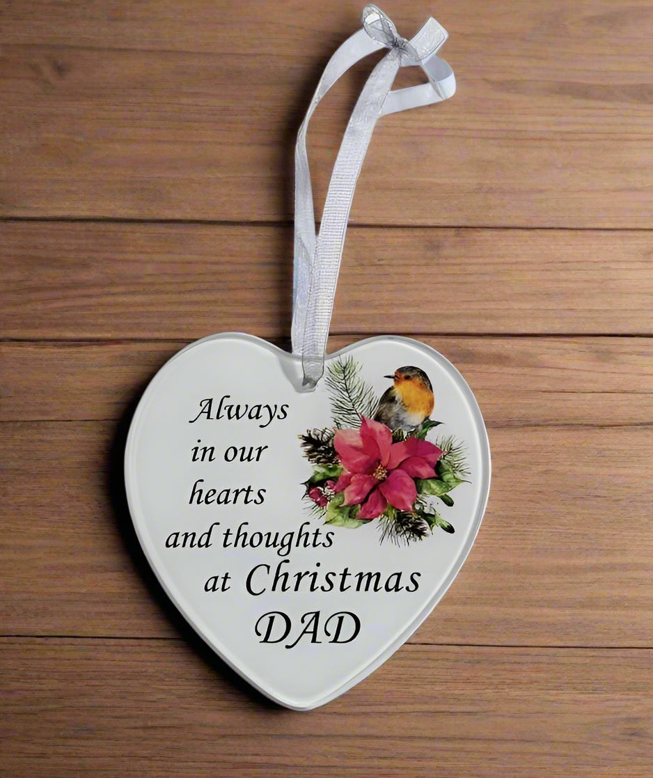A heart-shaped Christmas ornament with a white background, printed with the text 'Always in our hearts and thoughts at Christmas DAD' alongside a robin bird and red flowers, hanging from a white ribbon on a wooden background.