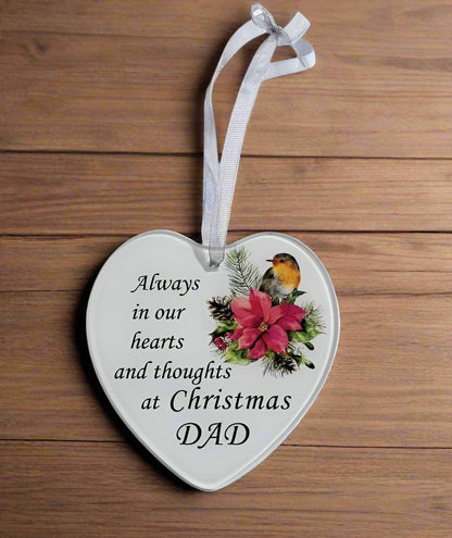 A heart-shaped Christmas ornament with a white background, printed with the text 'Always in our hearts and thoughts at Christmas DAD' alongside a robin bird and red flowers, hanging from a white ribbon on a wooden background.