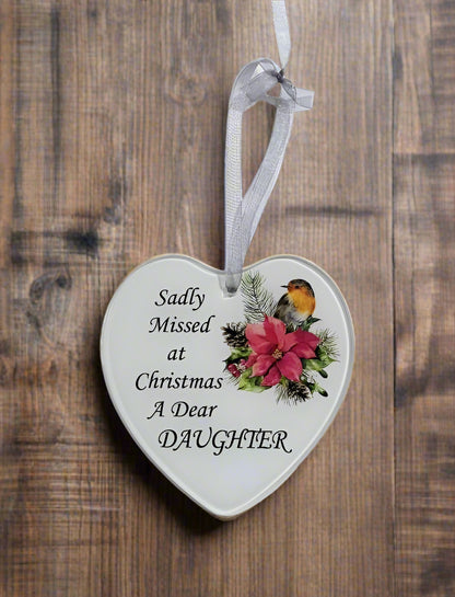 A heart-shaped white ceramic Christmas ornament hanging from a white ribbon, featuring the printed message 'Sadly Missed at Christmas A Dear DAUGHTER' alongside a festive illustration of a robin, poinsettia, and pinecones, set against a rustic wooden background.