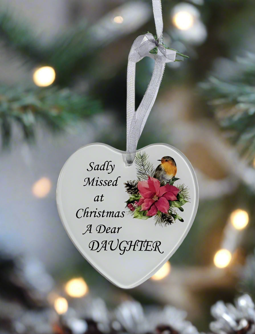A heart-shaped white Christmas ornament hanging from a white ribbon, printed with the text 'Sadly Missed at Christmas A Dear DAUGHTER' and decorated with a robin bird and pink poinsettia flower, set against a blurred Christmas tree background.