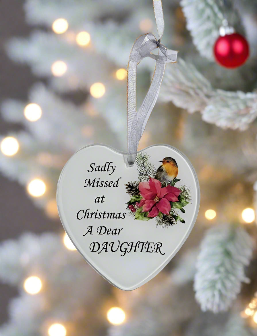 A heart-shaped white Christmas ornament hanging from a white ribbon, printed with the text 'Sadly Missed at Christmas A Dear DAUGHTER' and decorated with a robin bird and pink poinsettia flower, set against a blurred Christmas tree background with warm lights.