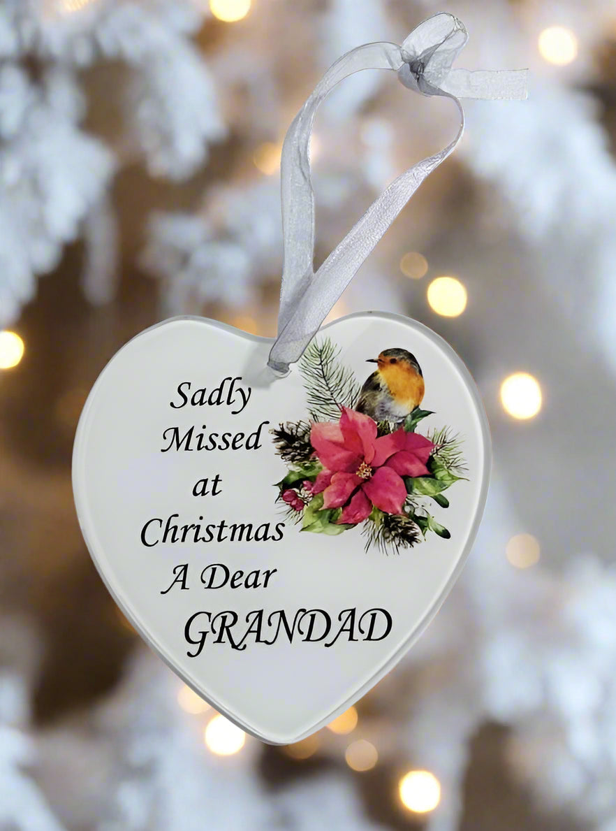 A heart-shaped white Christmas ornament with a silver ribbon hanger, printed with the text 'Sadly Missed at Christmas A Dear GRANDAD' alongside a robin bird and pink flowers, set against a blurred festive background.