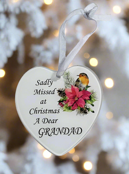 A heart-shaped white Christmas ornament with a silver ribbon hanger, printed with the text 'Sadly Missed at Christmas A Dear GRANDAD' alongside a robin bird and pink flowers, set against a blurred festive background.