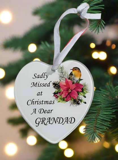 A heart-shaped white Christmas ornament hanging from a green pine tree branch, with a white ribbon loop. The ornament features black text reading 'Sadly Missed at Christmas A Dear GRANDAD' alongside an illustration of a robin bird and pink flowers.