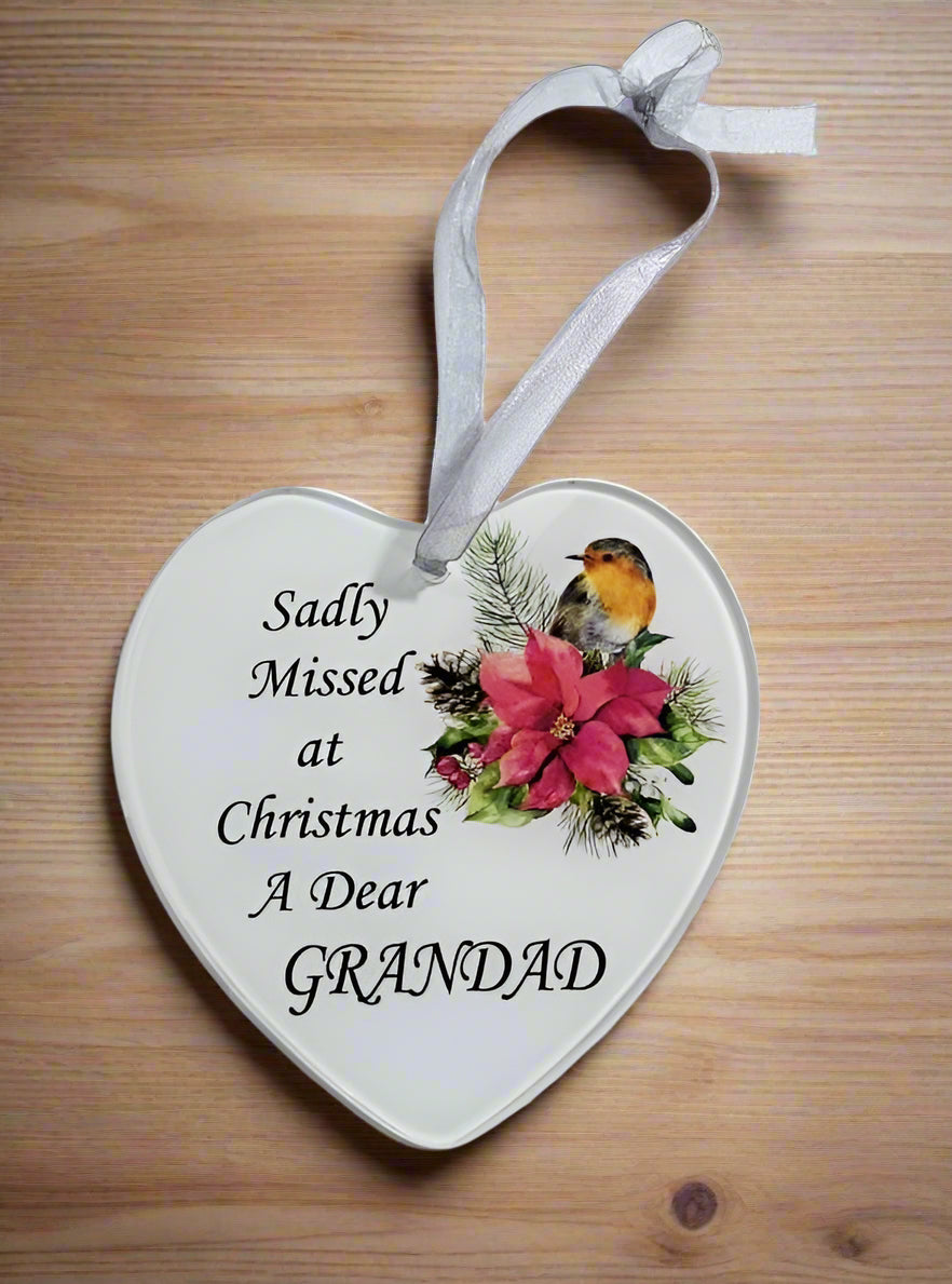 A heart-shaped white ceramic Christmas ornament hanging on a light wood surface. The ornament features the printed text 'Sadly Missed at Christmas A Dear GRANDAD' alongside an illustration of a robin bird and pink poinsettia flowers.