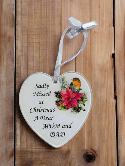 A heart-shaped ceramic Christmas ornament with a silver ribbon loop, featuring the printed text 'Sadly Missed at Christmas A Dear MUM and DAD' alongside a red poinsettia, green leaves, pinecones, and a small bird on a rustic wooden background.