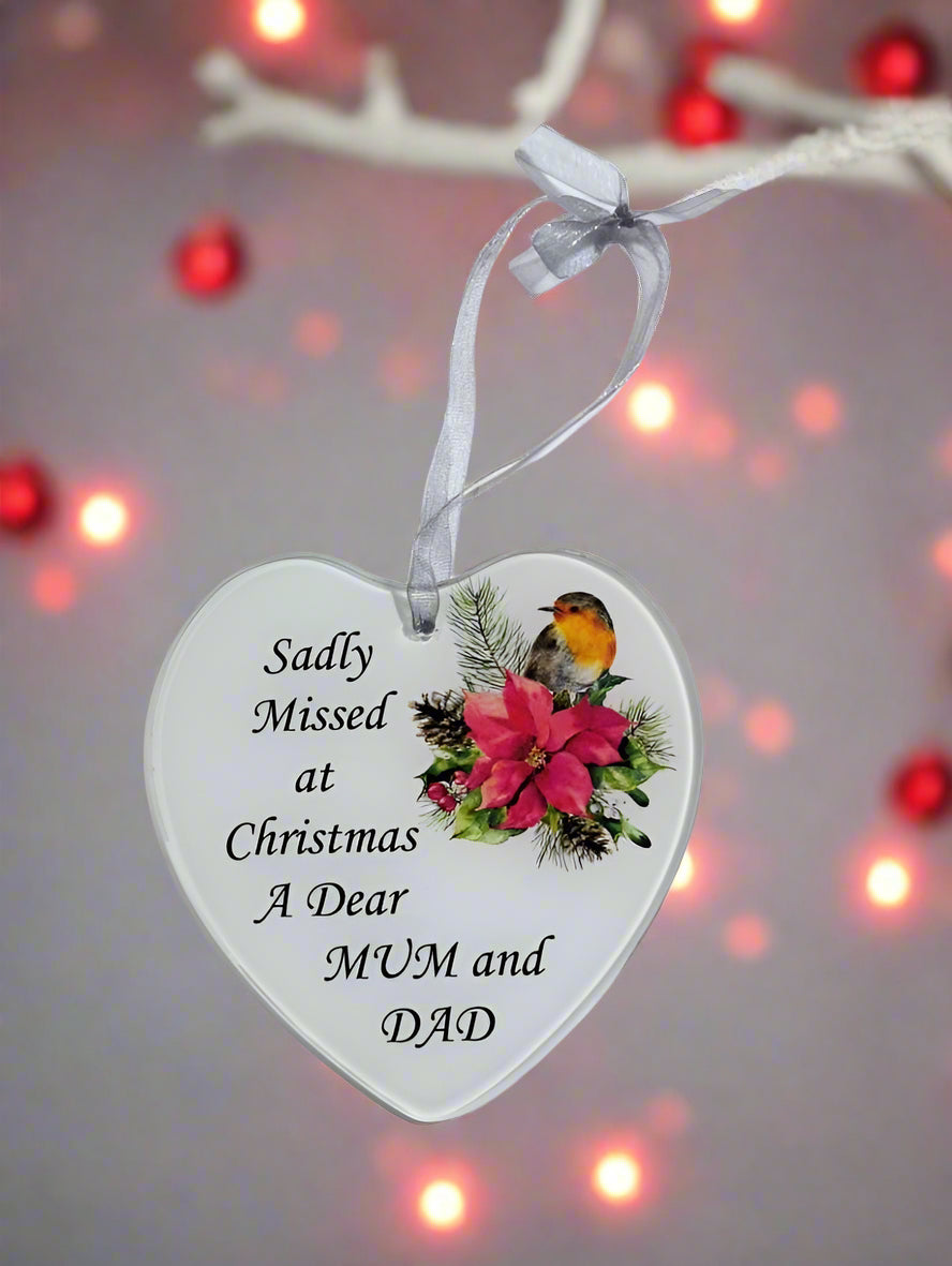 A heart-shaped white ceramic Christmas ornament hanging from a white ribbon, printed with the message 'Sadly Missed at Christmas A Dear MUM and DAD' alongside a robin bird and pink flower illustration, set against a blurred background with warm lights.