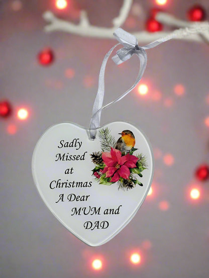 A heart-shaped white ceramic Christmas ornament hanging from a white ribbon, printed with the message 'Sadly Missed at Christmas A Dear MUM and DAD' alongside a robin bird and pink flower illustration, set against a blurred background with warm lights.