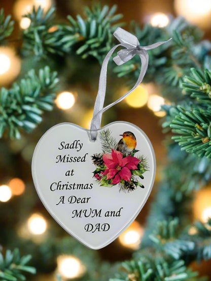 A heart-shaped white Christmas ornament with a gray ribbon, printed with the text 'Sadly Missed at Christmas A Dear MUM and DAD' and decorated with a robin bird and pink poinsettia flowers, hanging in front of a blurred Christmas tree with warm bokeh lights.