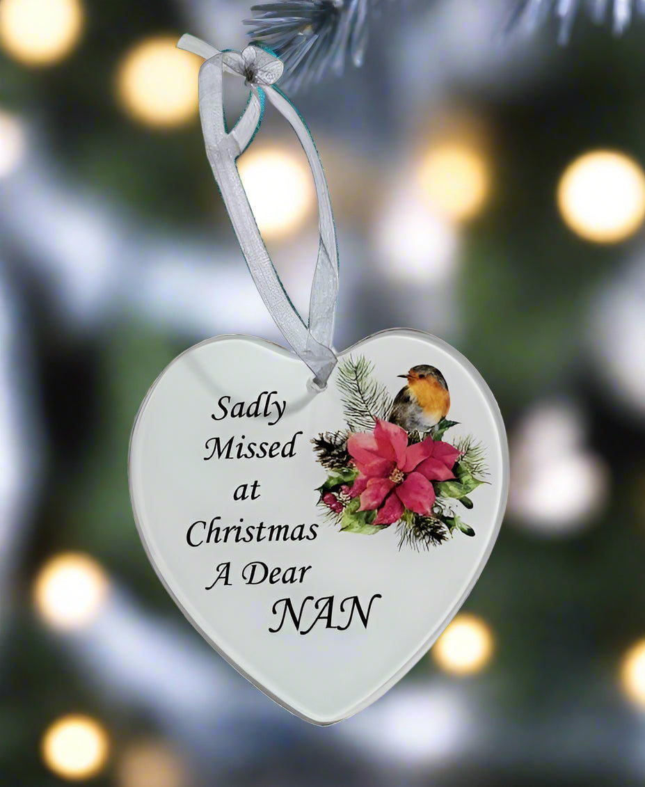 A heart-shaped white Christmas ornament hanging from a white ribbon, printed with the text 'Sadly Missed at Christmas A Dear NAN' alongside a robin bird and pink poinsettia illustration, set against a blurred Christmas tree background with warm lights.