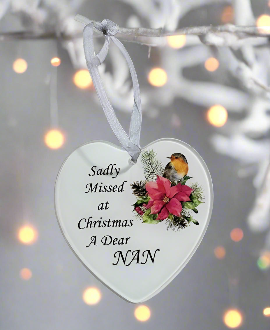A heart-shaped white Christmas ornament hanging from a white branch, printed with the message 'Sadly Missed at Christmas A Dear NAN' alongside an illustration of a robin bird, a pink poinsettia, and green foliage. The background features soft, out-of-focus warm bokeh lights.