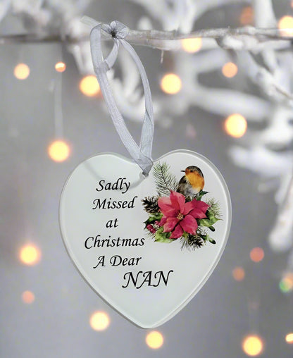 A heart-shaped white Christmas ornament hanging from a white branch, printed with the message 'Sadly Missed at Christmas A Dear NAN' alongside an illustration of a robin bird, a pink poinsettia, and green foliage. The background features soft, out-of-focus warm bokeh lights.