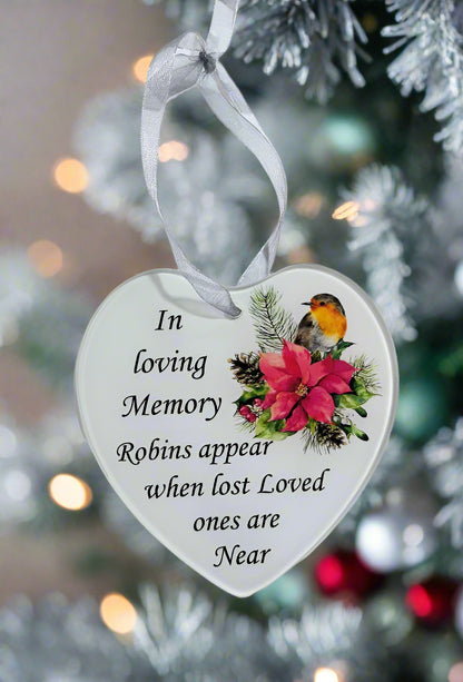 A heart-shaped white Christmas ornament hanging from a white ribbon, printed with the memorial message 'In loving Memory Robins appear when lost Loved ones are Near' and decorated with a robin bird and red flowers, set against a blurred Christmas tree background.