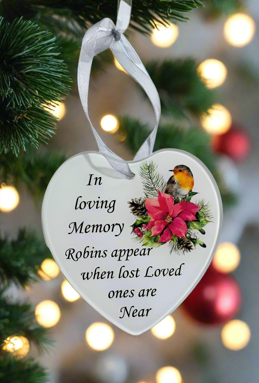 A heart-shaped white Christmas ornament hanging from a white ribbon on a Christmas tree. The ornament features black printed text reading 'In loving Memory Robins appear when lost Loved ones are Near' alongside an illustration of a robin bird and red flowers.