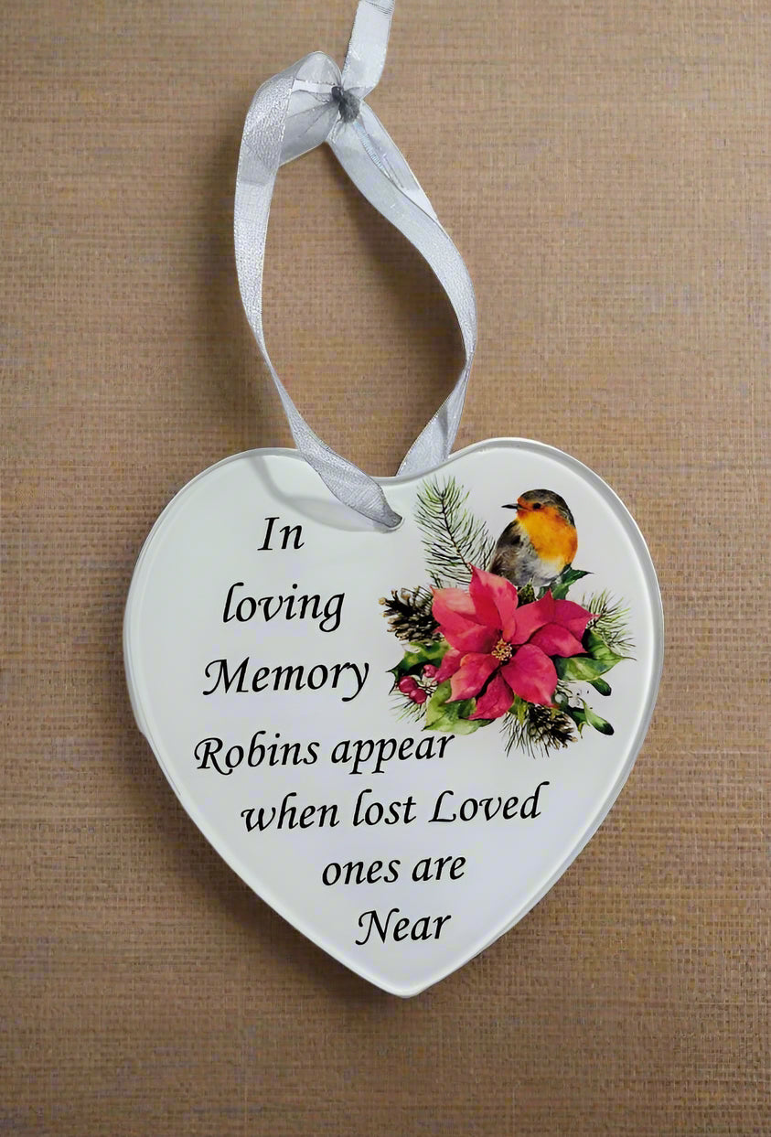 A white heart-shaped hanging memorial ornament with a white ribbon loop, printed with the phrase 'In loving Memory Robins appear when lost Loved ones are Near' and an illustration of a robin bird with pink flowers and green foliage, shown against a tan textured background.