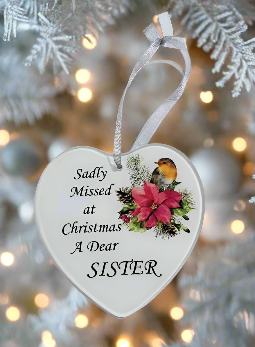 A heart-shaped white Christmas ornament hanging from a white ribbon, printed with the text 'Sadly Missed at Christmas A Dear SISTER' and decorated with a robin bird and pink poinsettias, set against a blurred white Christmas tree with warm lights.