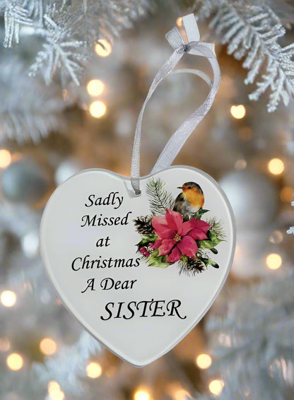 A heart-shaped white Christmas ornament hanging from a white ribbon, printed with the text 'Sadly Missed at Christmas A Dear SISTER' and decorated with a robin bird and pink poinsettias, set against a blurred white Christmas tree with warm lights.