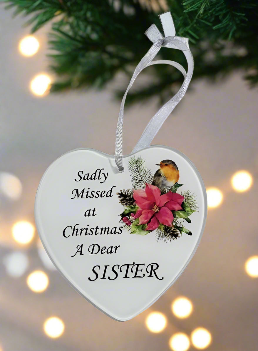 A heart-shaped white Christmas ornament hanging from a white ribbon, printed with the text 'Sadly Missed at Christmas A Dear SISTER' and decorated with a bird, pink flowers, and green foliage, set against a blurred holiday background with warm lights.