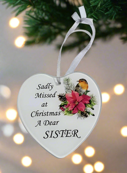 A heart-shaped white Christmas ornament hanging from a white ribbon, printed with the text 'Sadly Missed at Christmas A Dear SISTER' and decorated with a bird, pink flowers, and green foliage, set against a blurred holiday background with warm lights.