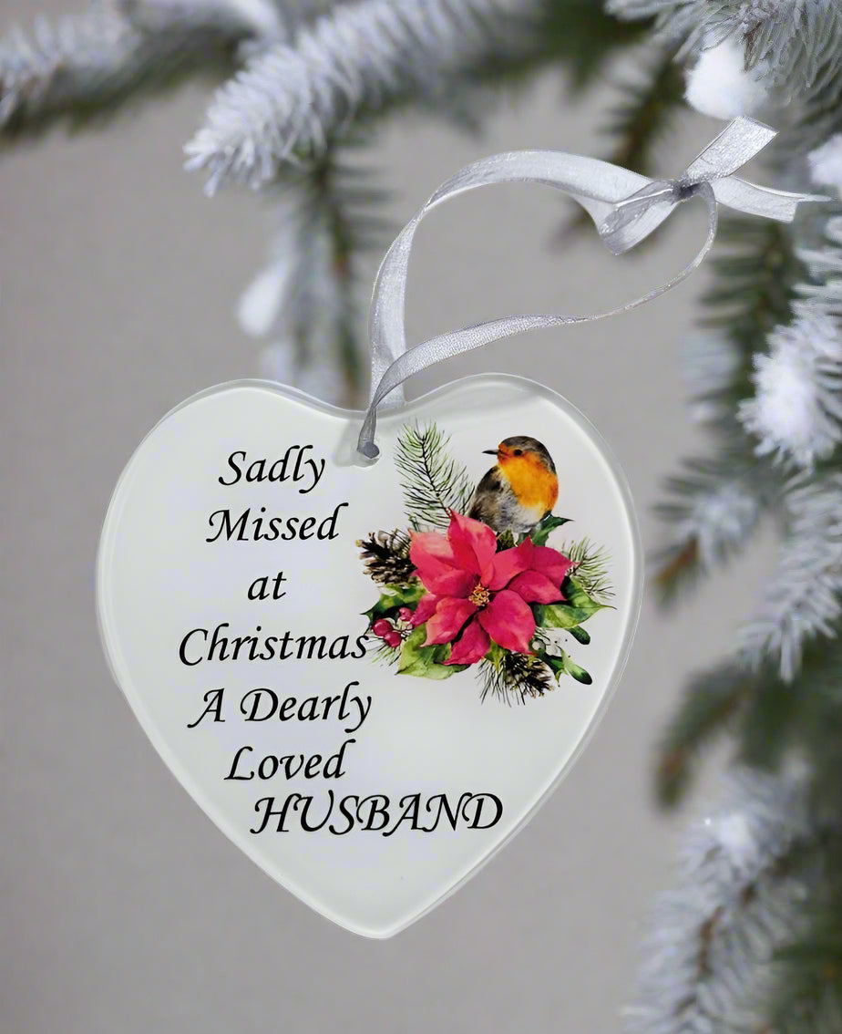 A white heart-shaped glass hanging decoration hanging on a Christmas tree with a poinsettia flower and a robin. The text on the decoration reads 'Sadly missed at Christmas a dearly loved Husband'.