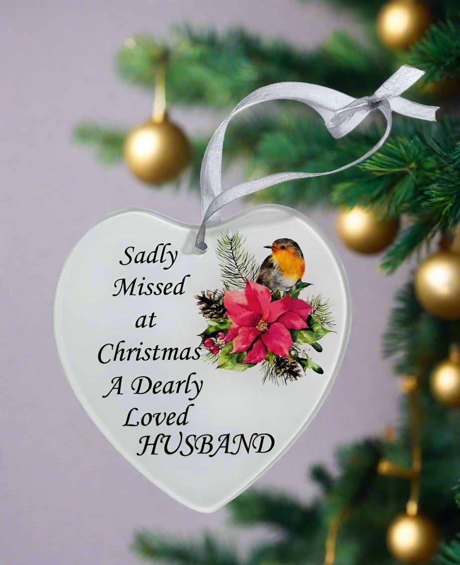 A white heart-shaped glass hanging decoration hanging on a Christmas tree with a poinsettia flower and a robin. The text on the decoration reads 'Sadly missed at Christmas a dearly loved Husband'.