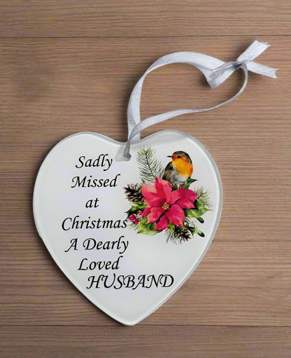 A white heart-shaped glass hanging decoration with a poinsettia flower and a robin. The text on the decoration reads 'Sadly missed at Christmas a dearly loved Husband'.