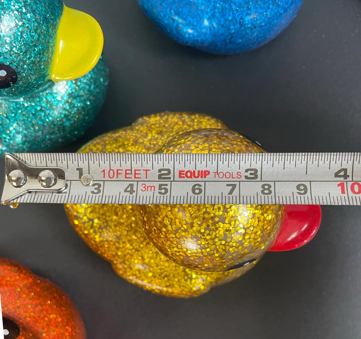 A close-up of a yellow glittery rubber duck with a red beak, shown with a tape measure across its body to indicate size. Other glittery duck toys in teal, blue, and orange are partially visible in the background on a dark surface.