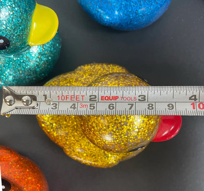 A close-up of a yellow glittery rubber duck with a red beak, shown with a tape measure across its body to indicate size. Other glittery duck toys in teal, blue, and orange are partially visible in the background on a dark surface.