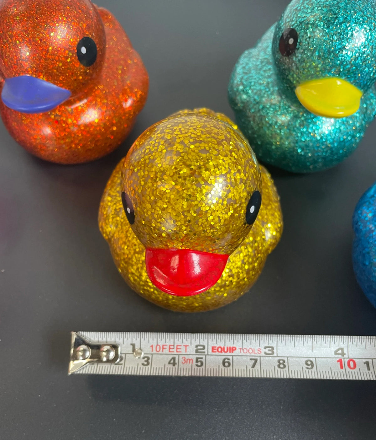 A glittery rubber duck toy in gold with a red beak sits on a dark surface, with a metal tape measure placed in front of it. Other glittery duck toys in orange, teal, and blue are partially visible in the background.