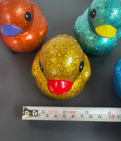 A glittery rubber duck toy in gold with a red beak sits on a dark surface, with a metal tape measure placed in front of it. Other glittery duck toys in orange, teal, and blue are partially visible in the background.