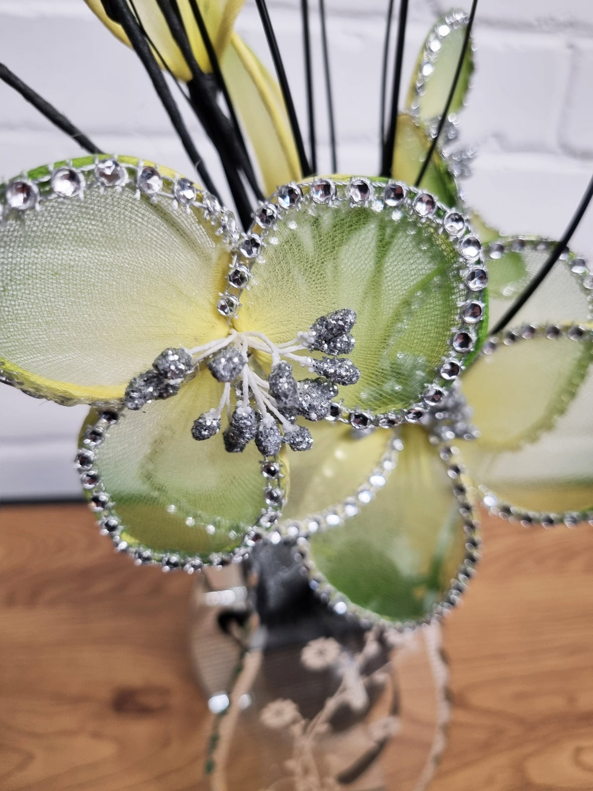 A decorative floral arrangement featuring large iridescent green and yellow petal-like pieces outlined with silver rhinestones, with black wire stems and a silver embellished center, displayed in a vase on a wooden surface.