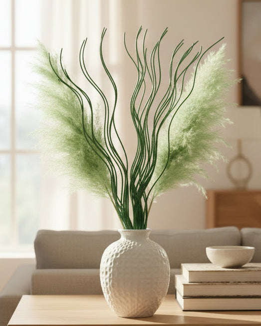 A decorative artificial pampas grass arrangement in a textured white ceramic vase, placed on a light wood table in a softly lit living room setting.