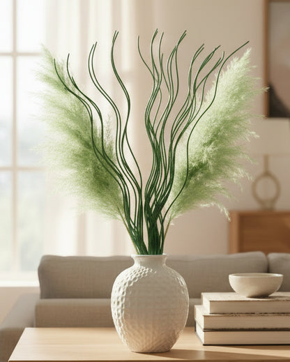 A decorative artificial pampas grass arrangement in a textured white ceramic vase, placed on a light wood table in a softly lit living room setting.