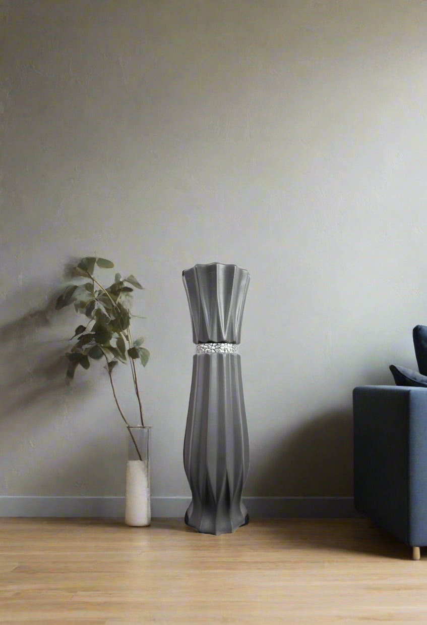 A modern gray pleated floor lamp stands on a light wood floor against a textured gray wall, styled next to a small white vase with a plant and a dark blue armchair.
