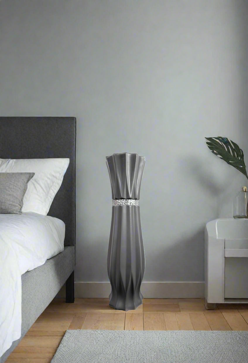 A tall, sculptural gray floor lamp with a flared top and a silver metallic band around the middle, standing in a modern bedroom next to a bed and a white side table.