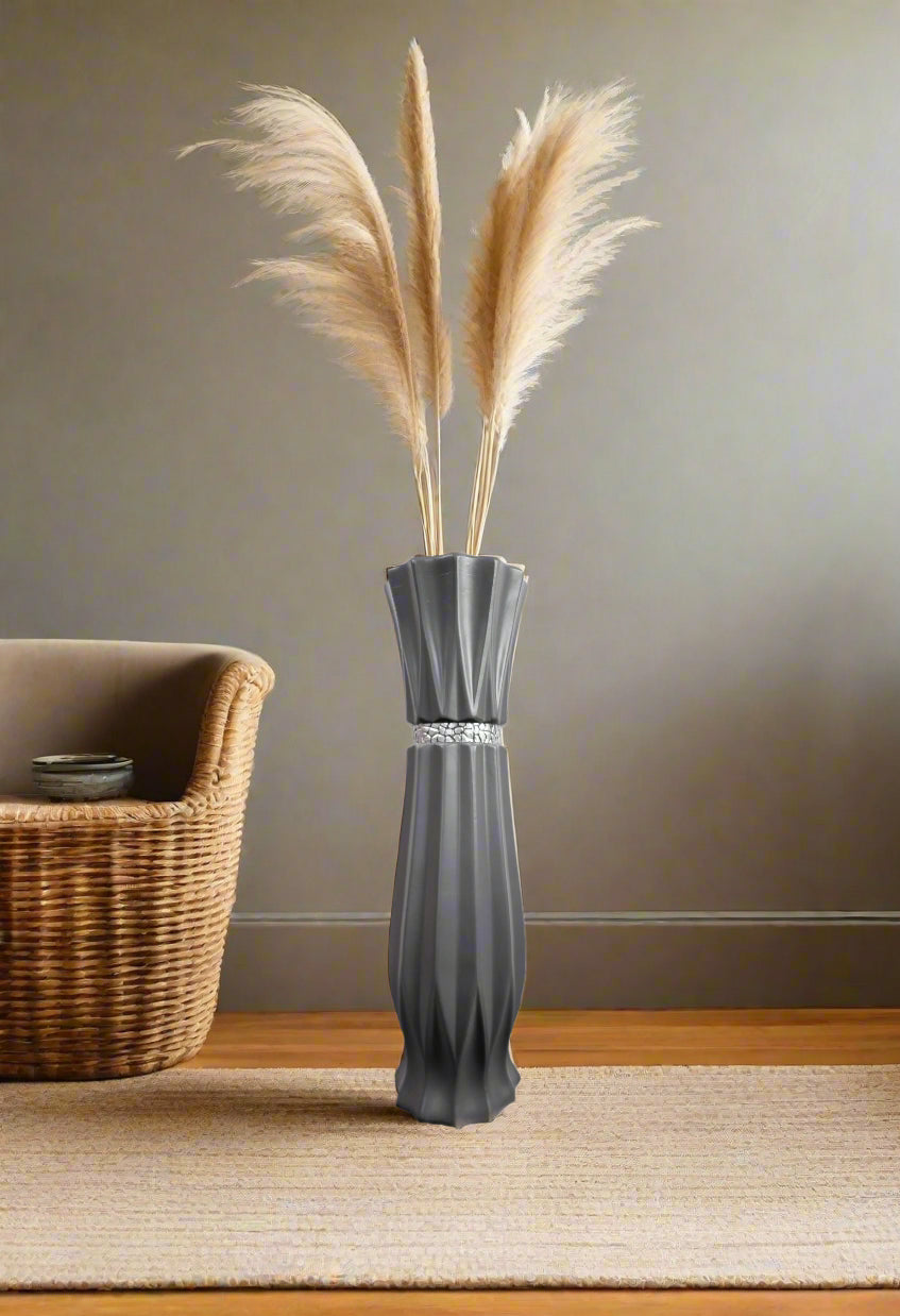 A tall, dark gray fluted ceramic vase with a silver metallic band at the neck, filled with dried pampas grass, set on a woven rug next to a wicker basket against a neutral gray wall.