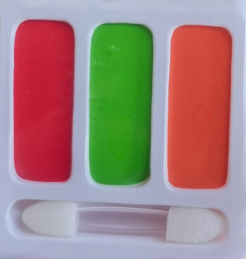 A makeup palette containing three rectangular pans of cream makeup in red, green, and orange, with a small dual-ended sponge applicator resting in the bottom slot.