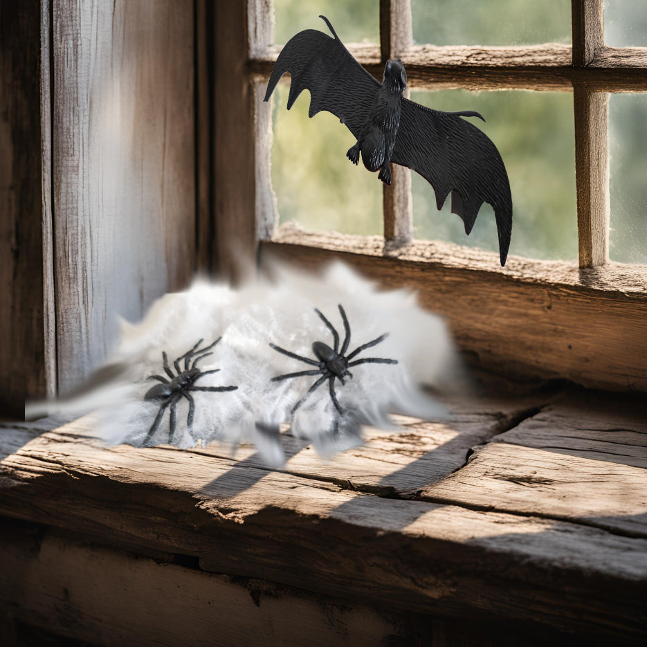 Halloween decoration set featuring black plastic spiders and a bat flying over white fake cobwebs arranged on a rustic wooden windowsill.