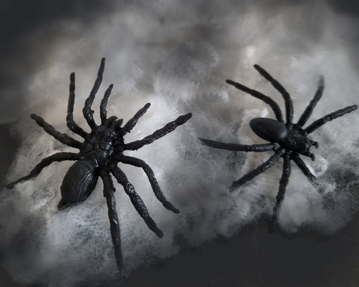 Two realistic black plastic toy tarantulas resting on white faux cobwebs against a dark, smoky background.