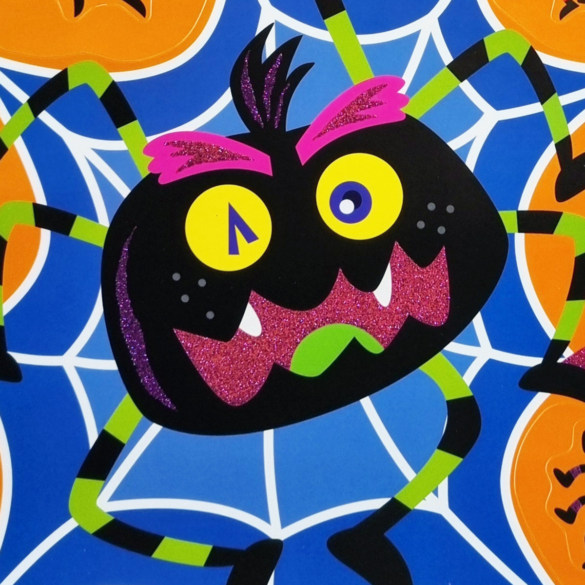 A colorful Halloween-style monster spider illustration on a blue spiderweb background with orange pumpkin shapes. The spider has a black body, bright yellow eyes, pink glittery eyebrows and mouth details, green and black striped legs, and a small green tongue.