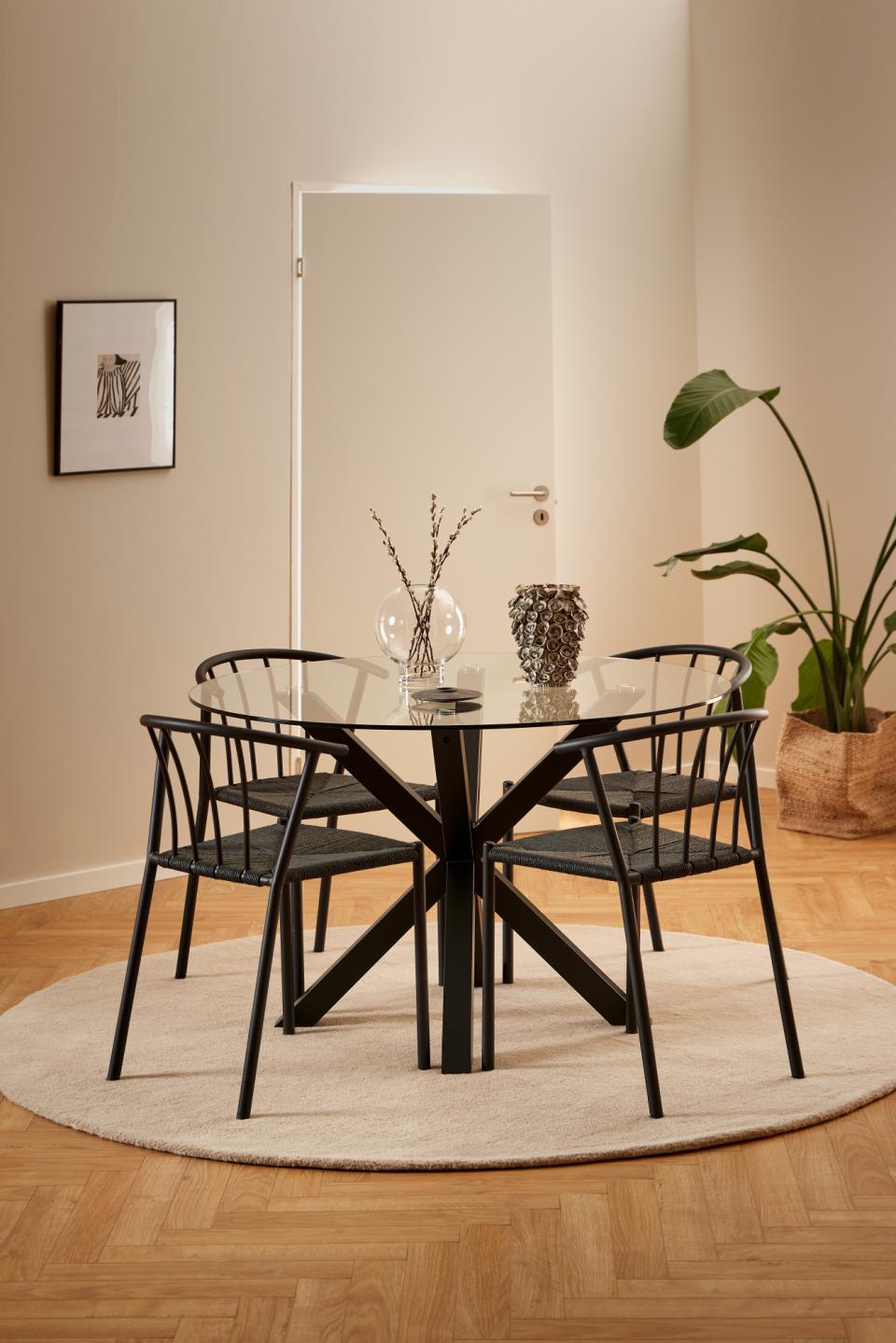 A modern dining set featuring a round glass-top table with a black metal X-shaped base and four matching black chairs with woven backs and seats, arranged on a round off-white rug in a warm, minimalist dining room.